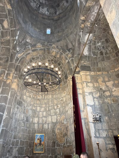 Stone vaulted ceiling, likely of a church or cathedral, dominates the view. A large, circular chandelier with a cross design hangs in the center, encircled by lights. To the right, a red banner or curtain runs along the wall. A small framed painting is below the banner. There is also a small window visible in the dome. The stonework is intricate and gives the space a historical feel.