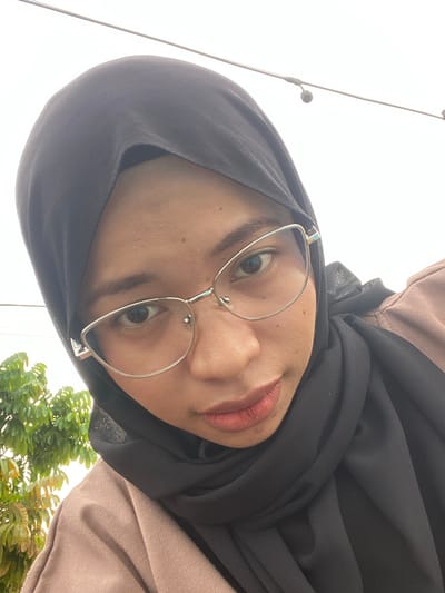 The media file shows a close-up, downward-angled selfie of a young woman wearing a black hijab and glasses. Her head is tilted, with her face filling most of the frame. She is wearing a light brown hooded top underneath the hijab. Her lips are slightly parted and appear to be painted with lipstick. The background is bright and out of focus, with a hint of green foliage on the right side, suggesting an outdoor setting. Two thin poles with small dark circular objects are visible on the left, possibly part of a street light or other outdoor fixture. The lighting suggests it is daytime, with bright, overcast conditions. The woman's expression is neutral. No text is visible in the image. The location context is Air Molek, Indonesia.