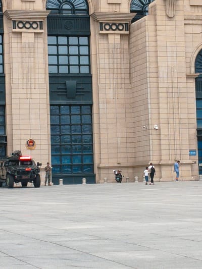 This image depicts a scene outside a large, tan stone building with prominent dark blue windows and architectural details. In the foreground, a military-style vehicle with flashing red lights is parked. A soldier in uniform stands to the right of the vehicle, facing the building. Further to the right, a family consisting of a woman, a young boy, and another person are walking away from the camera. A motorcycle is parked to the left of the family. The ground is paved with large, light-colored square tiles. The building appears to be a public or official structure, possibly a government building or a historical landmark. The overall impression is one of security and public presence, with the military vehicle and soldier suggesting a heightened level of vigilance. The architecture and signage, if any were visible and legible, might offer further clues to the specific location within Harbin, China.