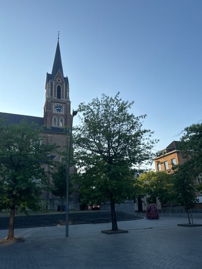 The image depicts a street view in Strombeek-Bever, Belgium. Dominating the scene is the prominent brick tower of the Church of St. Amandus and St. Amelberga, featuring a dark spire topped with a cross and a clock face with Roman numerals. The church is flanked by mature trees with green foliage. In the foreground, a paved square with a cobblestone pattern stretches across the lower portion of the image. To the right of the church, a modern brick building with windows is visible, partially obscured by trees. A large, dense bush or tree adorned with pink flowers is situated in the middle ground to the right. The sky is clear and blue. The scene appears to be captured during daylight, with the sun casting subtle shadows. No individuals are present in the frame.