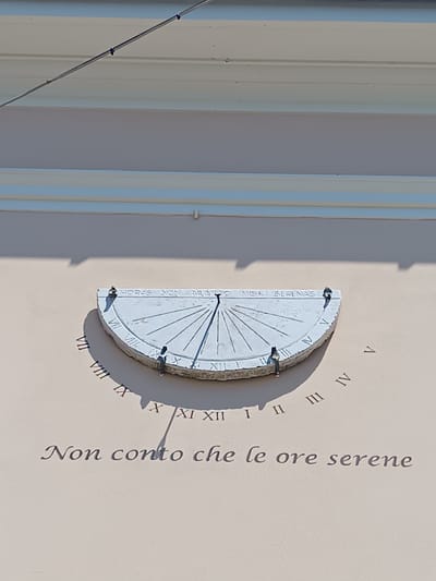 The image shows a sundial mounted on a light pink wall, likely part of a building in Mezzana Sant'Antonio, Italy. The sundial is a semi-circular stone structure with Roman numerals marking the hours. The gnomon casts a shadow indicating the approximate time. The Latin phrase "HORAS NON NUMERO NISI SERENAS" is inscribed on the dial. Below the sundial, the Italian phrase "Non conto che le ore serene" is written on the wall. A white trim runs along the top of the wall. A thin electrical wire runs above the trim.