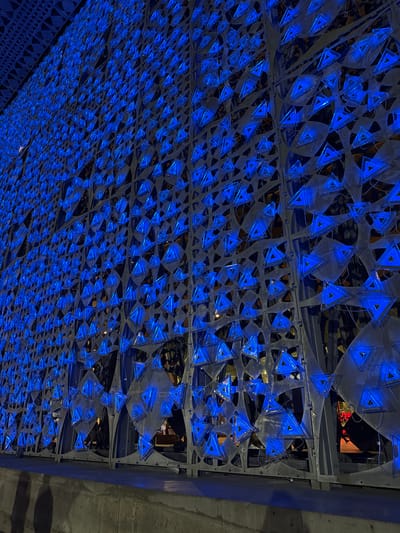 The image depicts an illuminated, triangular mesh structure on the exterior of a building in the city of Kruja, Albania. The structure is a large, dark metal grate with countless small, hollow triangles embedded within it. These triangles are illuminated from within by a bright, electric blue light, creating a vibrant and intricate pattern. The lighting casts a blue glow on the surrounding area and reflects off the metallic surfaces of the structure. In the foreground, a concrete ledge or sidewalk is visible, with the shadows of two people cast upon it. The background is dark and somewhat obscured, suggesting it is nighttime. The overall scene is modern and artistic, with the blue lights creating a striking visual effect against the dark backdrop.