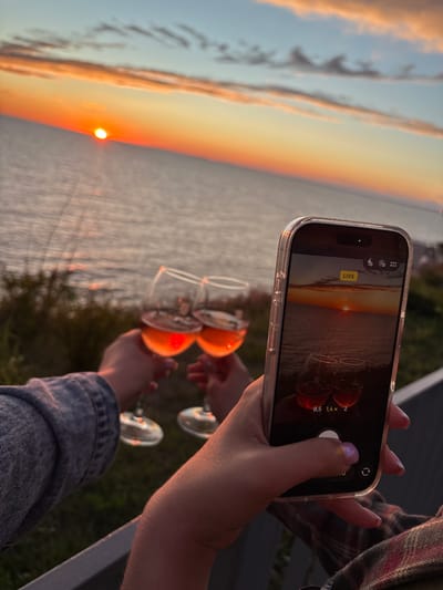 The image depicts a vibrant sunset over a vast body of water, likely a sea or large lake, consistent with the Vyshesteblievskaya region of Russia. The sun is positioned low on the horizon, casting brilliant orange and yellow light across the sky and reflecting on the water's surface. Faint clouds in the upper left also catch the warm glow.

In the foreground, a person's hand, wearing a plaid shirt, holds up a smartphone (an iPhone, identifiable by its interface and notch) to capture the scene. The phone's screen displays the sunset, intentionally composed to include two wine glasses in the lower part of the frame, mirroring the real-life setup. On the phone screen, the word "LIVE" is visible, alongside zoom options "0.5," "1.4x," and "2," and various camera mode icons.

Below the phone, another person's hands, clad in a denim jacket, hold two clear wine glasses. Each glass contains an orange-hued beverage, possibly rosé wine or an aperitif, held up towards the sunset. In the background, out of focus, two additional wine glasses can be vaguely seen.

The scene captures a moment of leisure and appreciation for nature, likely a social gathering or a romantic evening. The activity involves simultaneously enjoying and documenting the beautiful sunset with drinks, creating a composed photograph within a photograph. The time of day is clearly late evening, marking the end of the day with warm, tranquil light.