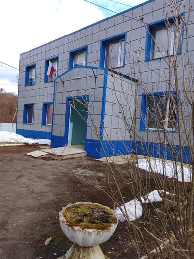 The image depicts a two-story, rectangular building with a gray facade and blue trim, likely located in Zhukov, Russia, as evidenced by the Russian flag displayed near the upper left window. The building has a simple, functional design with regularly spaced windows. A blue-trimmed doorway leads into the building. A security camera is visible above the entrance. In the foreground, there's an overgrown concrete planter filled with moss. Scattered patches of snow remain on the ground, suggesting a late winter or early spring season. Bare branches of a shrub are prominently featured in the right foreground, partially obscuring the view. The foreground also includes a paved area with visible cracks and discoloration. A grassy hill with leafless trees can be seen in the background.