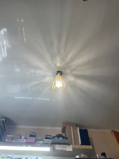 The image captures an indoor scene, likely a commercial or workspace, dominated by a brightly lit, ornate light fixture mounted on a light-colored, reflective ceiling or wall. The fixture, possibly a chandelier or decorative sconce, features a bright bulb surrounded by crystalline or beaded elements, casting strong, radial light patterns across the textured surface.

To the right of the frame, a wooden shelf or counter holds various objects, suggesting a storage or work area. These include several cardboard boxes, a dark laptop or electronic device, and a long fluorescent light fixture positioned vertically. The overall lighting is artificial and bright.

Visible text on various boxes and labels provides context:
*   "won Digital Multimeters"
*   "ΠΡΟΓΡΑ" (PROGRA)
*   "ФЕРРОЛИ" (FERROLI)
*   "BAX" (BAXI)
*   "MAIN 24-ECENTA"
*   "1 литр"
*   "NEVA LUX"
*   "VAILLANT" (a brand of heating, ventilation, and air conditioning equipment, prominently displayed on a sign as well)
*   "PHILIPS"
*   "Samsunq 132 10160 SAMSUNG SAMSUNG"
*   "WEUPASQ 732,491,41114"
*   "40%"
*   Handwritten notes include "по вопросам ТАРИФ" (on tariff issues) and another partially obscured Russian text.

The setting, with its mix of decorative lighting and practical, technical equipment, suggests an office, showroom, or workshop in Krayevsko-Armyanskoye, Russia. No people or activities are present.