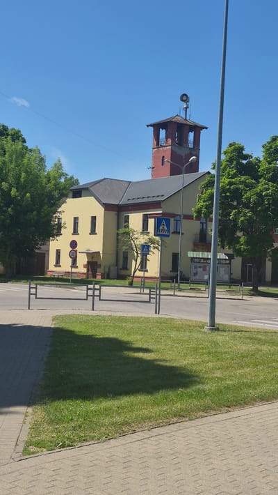 The image captures a view of the Preiļi Firefighter Station and its surrounding area in Preiļi, Latvia, on a clear, sunny day. The main subject is a two-story yellow building with a red tower, which is characteristic of a fire station, featuring an air raid siren at its apex. The building has a dark, sloped roof with visible ventilation grilles. In front of the building, there is a road with clear markings and pedestrian crossings, indicated by blue and white diamond-shaped signs. Several metal barriers are present along the sidewalk, which is paved with interlocking concrete stones. A strip of green grass borders the sidewalk and separates it from the road. In the background, lush green trees frame the scene, and a tall metal pole, likely a lamppost or part of a utility structure, stands prominently to the right of the building. No individuals are visible in the image.