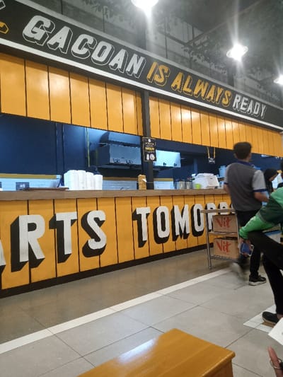 The image displays the interior of a Mie Gacoan restaurant in Nganjuk, Indonesia. A prominent service counter, faced with yellow panels and white 3D-effect lettering partially visible as "ARTS TOMORR", occupies the mid-ground. Above the counter, a black banner with white and yellow text states "GACOAN IS ALWAYS READY". Behind the counter, a dark blue wall section with yellow paneling conceals a food preparation area, where white stacked containers, stainless steel equipment, and exhaust hoods are visible through an opening. A small black vertical sign is present on the counter surface, and a "SELF SERVICE" sign on a chain is partially visible.
Two individuals are visible on the right side of the frame. One male, seen from behind, wears a dark grey collared shirt over a lighter grey t-shirt and dark trousers, appearing to move leftward. Adjacent to him, partially visible, is another person. In the immediate right foreground, the lower body of a person wearing dark trousers and black shoes, along with a portion of a green long-sleeved garment, is present, indicative of a delivery driver. A two-tiered metal trolley is situated between the first individual and the green-clad person, holding two brown cardboard boxes marked "Vit".
The floor consists of light grey tiles with a white demarcation line. A light brown wooden tabletop is partially visible in the lower foreground. The area is brightly illuminated by multiple ceiling-mounted circular light fixtures, revealing an exposed ceiling structure.