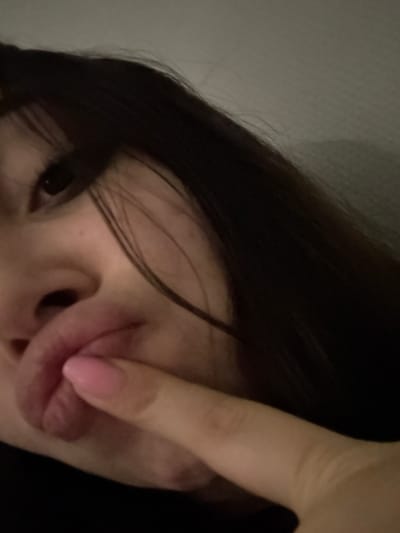 A selfie shows a person with dark hair, fair skin, and an eye visible. A hand with a pink nail touches their lips, partially obscuring their face. The angle of the photo is tilted, with the person's head positioned in the upper left corner of the frame. The background appears to be a light wall.