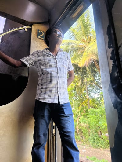 A man stands in a train carriage, looking out of an open doorway at lush greenery and palm trees, indicative of Aluthgama, Sri Lanka. The man, wearing a checkered shirt and dark jeans, has his left arm resting on the interior of the carriage and his right hand behind his back. He is looking upwards and to the right, with sunglasses on. A prominent number '2' is visible on the wall behind him, likely indicating a seat or carriage number. The lighting suggests it is daytime, with sunlight filtering through the vegetation. The scene captures a moment of observation from within the moving train.