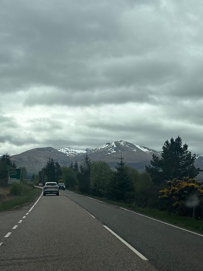 A view of a road in Spean Bridge, United Kingdom. The road stretches into the distance, with several cars traveling along it. To the left, a dramatic mountain range rises into a cloudy sky, with patches of snow clinging to the higher elevations. The roadside features trees and a signpost.
