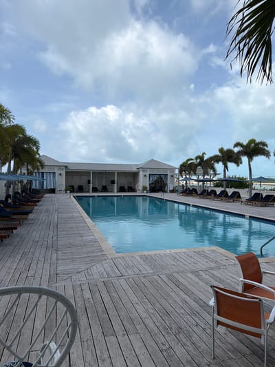 The image displays an outdoor swimming pool area in San Pedro Town, Belize, under an overcast sky with scattered clouds. No individuals are present. The foreground is dominated by a light-colored, weathered wooden deck, exhibiting planks laid in varying directions. A segment of a white wicker chair is visible in the lower-left, while two chairs with orange fabric seats and white frames occupy the lower-right. A clear blue rectangular swimming pool, bordered by light coping, centrally divides the midground. To the left of the pool, a row of approximately ten dark lounge chairs with dark blue cushions, each beneath a dark umbrella, extends along the wooden deck. Behind this arrangement, a long, white single-story building with a light gray gabled roof and white siding features several arched entrances, some adorned with blue and white striped awnings or curtains. Additional lounge seating is visible under the building's covered sections. To the right of the pool, another line of dark lounge chairs and umbrellas is positioned on the deck, backed by a white fence and a sequence of tall palm trees. In the background, beyond the fence and palm trees, a calm body of water is visible beneath the cloudy sky.