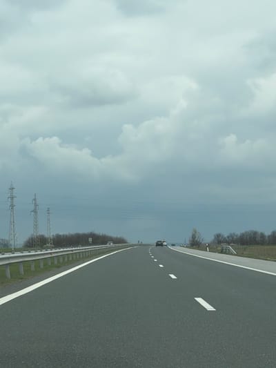 The image shows a view of a highway. The highway is made of asphalt, with white lane markings. There is a guardrail on the left side of the road. Beyond the guardrail is vegetation and power lines. In the distance, cars can be seen on the highway. The sky is cloudy. There is no evidence to suggest this is Vodenichane, Bulgaria.