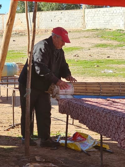 An elderly man in a red cap and dark clothing sits outdoors near a table draped with patterned fabric. He appears to be working with a white bucket. The backdrop features a brick wall, green grass, and a bamboo-like structure. A plastic bag sits on the ground by the table. The scene is bathed in sunlight, casting shadows on the ground. A partially visible wooden structure frames the foreground.