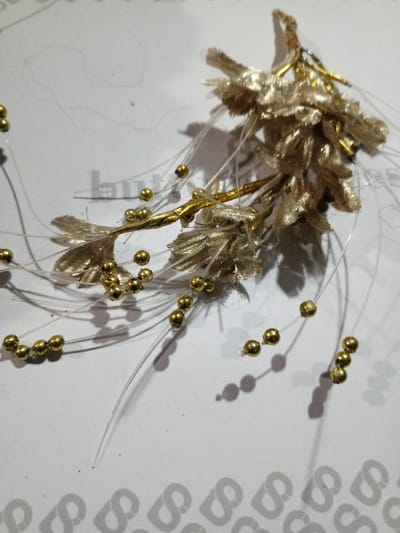 A golden floral hair accessory lies on a white surface patterned with faint gray logos. The accessory features fabric-like petals arranged in two clusters. Thin, clear filaments extend outwards, each punctuated with a small, golden bead. The light source casts shadows of both the floral element and the beaded filaments.
