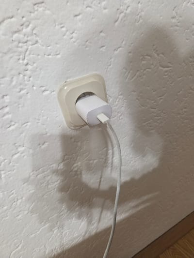 The scene depicts an interior wall in Negorci, North Macedonia. The wall is white and features a rough, textured surface. Mounted on this wall, positioned slightly diagonally, is a light beige, square-shaped electrical outlet of a European standard (e.g., Type F, Schuko). A white, compact power adapter, consistent with a mobile device charger, is plugged into this outlet. A light gray, braided cable extends downwards from the adapter. Shadows are cast on the wall, indicating an overhead or upper-right light source. A strip of light-colored wooden flooring is visible at the bottom right edge of the frame. No individuals, direct actions, or interactions are present in the image.