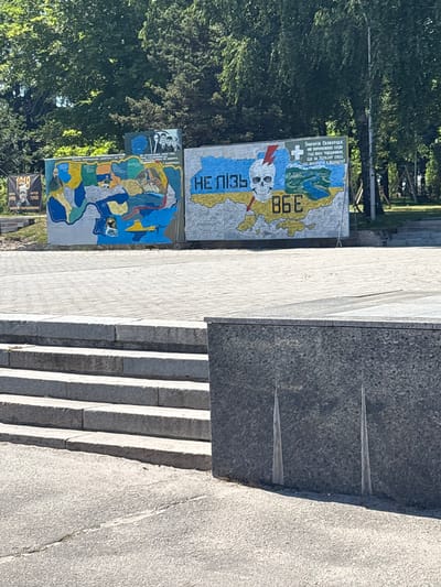 The image shows a mural on a wall, likely in Zmiiv, Ukraine, given the map of Ukraine depicted and the Ukrainian text. The mural features a stylized map of Ukraine with various regions highlighted in different colors. To the left of the map, there is a black banner with "1918" and an image of a man's face. Above the map, a quote from Hryhorii Skovoroda is visible, accompanied by a Christian cross and what appears to be a representation of nature. To the right of the map, the word "НЕПІЗЬ" (NEPIZ, meaning "never") is prominently displayed next to a skull with a lightning bolt through it. Below this, the Cyrillic letters "ВБЕ" (VBE) are visible. The mural is situated outdoors, with trees and foliage in the background. In the foreground, there are concrete steps and a paved area, suggesting a public space or monument.