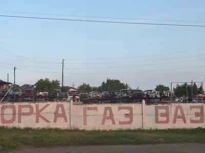 This media file depicts an outdoor scene at what appears to be a car junkyard or recycling facility in Beryozovka, Russia. The foreground features a low concrete wall with large, faded red painted letters spelling out "OPKA ГАЗ ВАЗ". These are likely abbreviations or names related to Russian car manufacturers, such as GAZ and VAZ.

Behind the wall, a collection of old, dismantled, and rusted car bodies are piled high on metal racks. These vehicles are the primary objects in the scene, showcasing a variety of makes and models in a state of disrepair, with many stripped of their parts. In the background, trees and some residential buildings are visible, suggesting the facility is located on the outskirts of a populated area.

The sky is a pale blue with a few wispy clouds, indicating it is daytime, likely late afternoon or early evening given the soft light. Several power lines crisscross the upper portion of the image, supported by wooden and metal poles. The overall impression is one of abandonment and decay, with the weathered appearance of the wall and the derelict vehicles contributing to a somber atmosphere. There are no people visible in the image. The presence of the car parts and the lettering on the wall clearly identify the location as a place for automotive salvage or disposal.