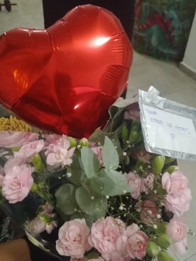 A red heart-shaped balloon sits beside a bouquet of pink carnations and baby's breath wrapped in clear cellophane. A small white card is attached to the bouquet, displaying handwritten words and a heart symbol. The background includes a glimpse of a patterned surface. The overall impression is a gift arrangement intended to convey love or affection.