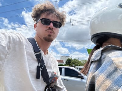 A man and a woman are on a motorbike in Nonthaburi City Municipality, Thailand. The man is in the foreground, wearing sunglasses and a white t-shirt, with curly hair and a goatee. He is making a pouty face. He has earbuds in and a bag slung across his chest. The woman is behind him, wearing a white helmet and a blue and tan plaid shirt. They are riding on a street with power lines overhead and some buildings and trees in the background. A silver car is visible to the right of the man. The sky is blue with some white clouds.