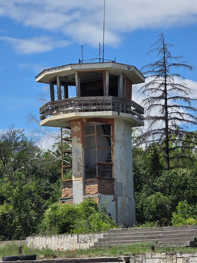 This is an exterior, full-shot, eye-level photograph of a derelict control tower located in Akatsievo, Bulgaria. The tower is octagonal in shape and appears to be made of concrete and brick. The windows are broken, and the paint is peeling, suggesting it has been abandoned for some time. A wooden balcony encircles the upper section of the tower. In the foreground, there are dilapidated concrete stairs and what appear to be remnants of a seating area. The tower is surrounded by lush green trees and vegetation, with a single, tall, dead tree to the right. The sky above is blue with scattered white clouds. There are no people or animals visible in the image. The scene conveys a sense of decay and abandonment within a natural setting.