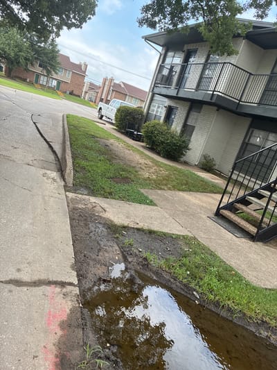 A low-angle, outdoor, eye-level shot shows a damaged sidewalk and a muddy puddle in front of a brick apartment building on a cloudy day. The sidewalk is cracked and tilted towards the street, with a muddy area filled with murky water and debris. Green grass lines the edge of the sidewalk and the apartment building. The apartment building has light-colored brick, dark balconies with metal railings, and several windows. A white pickup truck is parked on the street in front of a row of townhouses in the background. A black metal staircase with carpeted steps leads up to one of the apartment entrances. The overall scene suggests a neglected or poorly maintained urban environment.