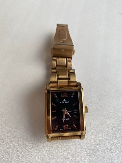 A gold-colored rectangular wristwatch with a black face lies on a white, textured surface. The watch has a metal band. The face displays the number 12 at the top, and the word "VECTOR" with a small triangular logo below it. At the bottom of the face, "WR 30M" is visible, as well as "JAPAN" and "MOVT". The watch is not in a city, but on a flat surface in an indoor setting.