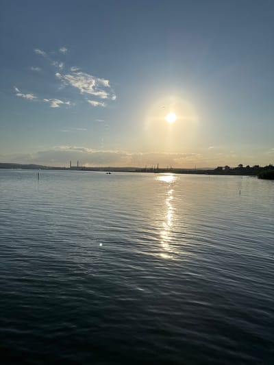 A wide-angle, eye-level, outdoor shot depicts a serene body of water under a bright, sunny sky. The sun is positioned in the upper center of the frame, casting a golden reflection across the rippling surface of the water. The sky is a clear blue with a few wispy white clouds scattered across it. In the distance, a long, low-lying shoreline is visible, featuring a collection of industrial buildings with tall chimneys, as well as some scattered trees and structures. The water appears calm, with gentle waves creating a textured pattern that leads the eye towards the distant horizon. The overall impression is one of tranquility and natural beauty, with a hint of industrial presence on the periphery. The location appears to be the Kazashko Reservoir in Bulgaria.