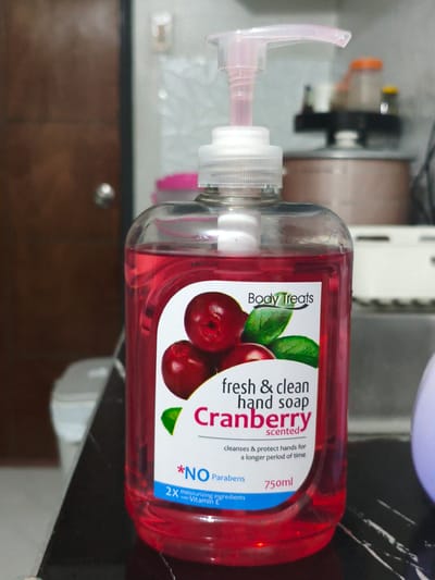In Cavite City, Philippines, a clear plastic bottle of "Body Treats" cranberry-scented hand soap stands on a dark, reflective surface. The bottle is filled with a vibrant red liquid and features a white label with images of cranberries and green leaves. Text on the label reads "fresh & clean hand soap," "cranberry scented," and "cleanses & protect hands for a longer period of time." Below this, it states "*NO Parabens" and indicates "2X moisturizing ingredients with Vitamin E" and a volume of "750ml." The background is blurred, showing hints of a wooden door on the left and a cluttered shelf with various objects, including containers with orange and yellow lids, and a beige appliance on the right. The lighting appears to be artificial, casting a slight glow on the bottle.