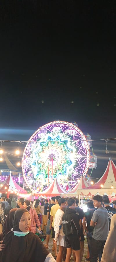 The image depicts a nighttime scene at what appears to be a local fair or carnival in Banding Agung, Indonesia. The central and most prominent feature is a large, brightly lit Ferris wheel, adorned with an intricate pattern of colorful lights in shades of purple, green, and white, creating a star-like or floral design. The wheel has several gondolas, some of which are visible.
In the foreground and middle ground, a crowd of people is gathered. They are standing, milling about, and some appear to be in line or observing the attractions. The people are dressed in casual attire. Several individuals are wearing hijabs, suggesting a culturally diverse or predominantly Muslim population. One woman in the immediate foreground is wearing a black hijab and a light blue surgical mask around her neck.
The environment includes tents with red and white striped awnings, and strings of yellow lights are strung across the area, illuminating the scene. The ground appears to be uneven and possibly muddy or dirt. In the background, the sky is dark, and there are several small, bright objects that could be sky lanterns. The overall atmosphere is festive and lively, characteristic of a community event held after dark. The scene suggests a popular gathering place in the town of Banding Agung.