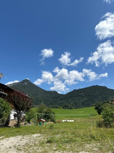 This image depicts a rural landscape in Bischofswiesen, Germany. In the foreground, a gravel path leads to a grassy area. To the left, a portion of a wooden house with a satellite dish is visible, along with a stack of firewood. A small, green and red playhouse stands in the middle ground, with a swing set nearby. Further back, a green field is dotted with several white hay bales and a red tractor. The background is dominated by a large, tree-covered mountain range under a bright blue sky with scattered white clouds.