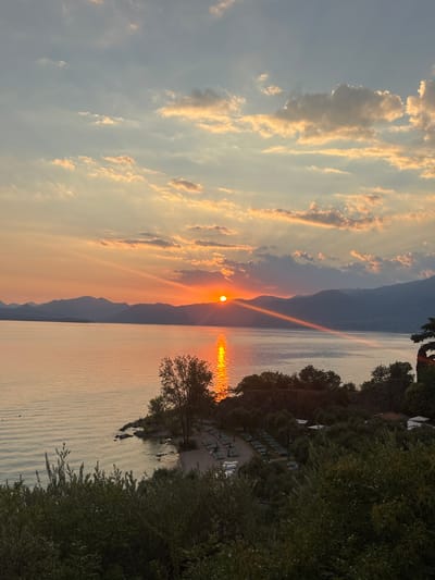 This is a photograph of a sunset over Lake Garda, Italy. The sun is low on the horizon, casting a warm golden light across the water. The sky is filled with dramatic clouds, tinged with orange and pink. In the foreground, lush green trees and foliage frame the scene, leading down to a small beach with rows of empty lounge chairs. Distant mountains form a silhouetted backdrop across the lake. The overall atmosphere is serene and picturesque.
