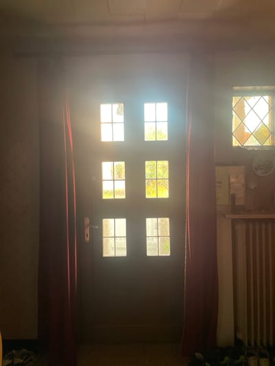 The image depicts an interior view of a doorway with multiple windows, illuminated by bright sunlight streaming through. The doorway is dark wood and features a brass handle. Above the doorway, there are two sets of windows: the top set has four panes, and the bottom set also has four panes. To the right of the doorway, a diamond-shaped window is visible, also letting in sunlight. Red curtains hang on either side of the doorway. In the background, a white radiator can be seen. The scene is lit by the intense sunlight, creating lens flare and partially obscuring details. The location is specified as Gerstheim, France.