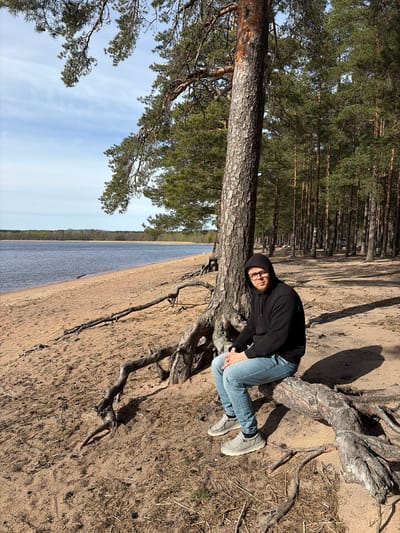 A person is lying at the base of a tree, on a sandy area near a body of water in Glubokoe, Russia. The person is wearing a black hooded sweatshirt, blue jeans, and light gray shoes. The tree is a pine, with exposed roots extending onto the sandy ground. Behind the tree, there is a dense pine forest. To the left of the tree, a body of water stretches to the horizon under a blue sky with some clouds. The scene is bathed in sunlight.