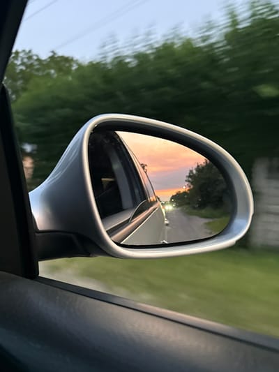 This media file captures a reflection in a car's side-view mirror, looking backward from the perspective of a moving vehicle. The mirror, a sleek silver color, is a prominent object in the foreground. Within the mirror's reflection, a sunset sky is visible, painted with hues of orange, pink, and yellow. This warm sunset contrasts with the cooler tones of the sky on the left side of the overall image.
The reflection also shows a road stretching into the distance, illuminated by streetlights. The road appears to be asphalt, and there are hints of other vehicles or light sources further down. Trees with green foliage line the sides of the road, blurred due to the motion of the car. In the background of the overall image, outside the mirror, the same blurred green trees and a sliver of pale sky are visible.
The time of day is clearly sunset, as indicated by the sky's colors and the streetlights becoming illuminated. The weather appears clear and calm, suitable for driving. The scene suggests a journey through a semi-rural or suburban area, likely in Petrushky, Ukraine, given the location context. There is a sense of movement and perhaps a moment of peaceful observation during travel. No text is visible in the media file.