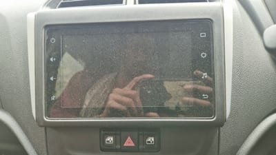 The image shows a close-up view of a car's infotainment system. The screen is covered in dust, obscuring much of its detail. On the left side of the screen, there are icons for power, volume up, volume down, and mute. On the right side, icons for a home screen, settings, music, and a return function are visible. Below the screen, there are three buttons: two with a window icon, presumably for power windows, and a central button with a red triangle and hazard lights symbol. The surrounding car interior is textured grey plastic. The image is taken from a slightly downward angle, showing the dashboard. There are reflections on the dusty screen, including what appears to be a person's hand and arm. No specific location in India is discernible from the image.