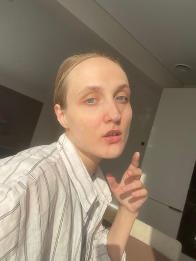 A light-skinned person with light hair is posing indoors, wearing a white and gray striped collared shirt. The person's face is prominently featured, revealing blue eyes and pink lips. The person's right hand is raised, fingers slightly spread. The interior space includes light-colored walls and cabinets. The photograph may have been taken near Sepych, Russia, but there's not enough information in the image to confirm this.