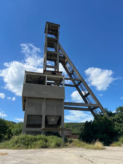 The image depicts a derelict industrial structure, likely a former processing plant or conveyor system, under a clear blue sky dotted with white clouds. The structure is made of concrete and appears to be a skeletal frame with open sections, suggesting it's abandoned or unfinished. A long, inclined conveyor belt structure dominates the scene, supported by a series of concrete pillars. Below and to the right, there are more concrete sections forming what might have been a processing or storage area.
The surrounding environment is lush with green trees and vegetation, indicating a rural or semi-rural setting. A dirt or gravel road runs along the right side of the frame, adjacent to the overgrown greenery. In the middle ground, partially obscured by trees, a small brick building with a few windows is visible, adding to the sense of a disused industrial site.
The lighting suggests it is daytime, likely mid-morning or afternoon, given the bright sunlight and clear sky. There is no activity or people visible in the image, emphasizing the sense of abandonment and stillness. The overall mood is one of decay and the passage of time, juxtaposed with the vibrant natural elements. No text is clearly legible in the image.