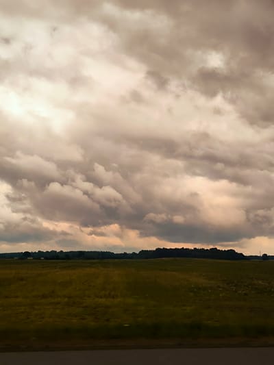 The image depicts a vast, open field under a dramatic, cloud-filled sky. The sky is a mix of light and dark gray clouds, with patches of warm, golden light breaking through, suggesting either sunrise or sunset. The field is covered in dry grass, tinged with yellow and brown, indicating it might be late summer or autumn. In the distance, a dark line of trees forms a horizon, behind which the sky is lighter. The bottom of the image shows a dark, blurry band that could be a road or a barrier, suggesting the photo was taken from a moving vehicle. There are no people or specific structures visible in the landscape. The scene conveys a sense of natural expanse and potentially impending weather change due to the stormy sky. The location is identified as Dunte, Latvia.