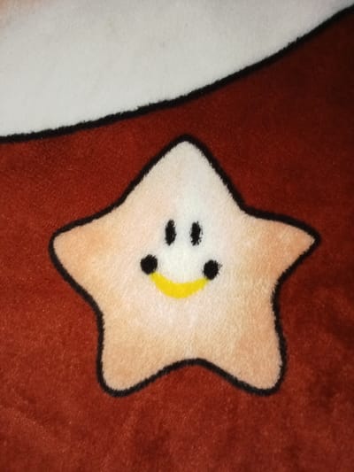 The image displays a close-up of a textile pattern in Ciputri, Indonesia. The primary subject is a five-pointed star, colored light beige with a distinct black outline. This star features a stylized cartoon face, detailed with two small black dots positioned above two larger black dots, and a curved yellow line below, forming a smile. The star is situated on a textured, reddish-brown fabric background. A segment of a white, black-outlined shape curves into the upper left portion of the frame, indicating it is part of a larger, multi-patterned textile item. No human figures, actions, or interactions are present.