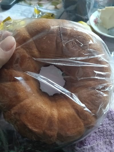 In the city of Altinkul, Uzbekistan, a person's hand, with manicured fingernails, is holding a ring-shaped cake wrapped in clear plastic. The cake is golden brown and appears to be a pound cake or similar dense confection, with fluted sides. The plastic wrap is slightly crinkled, obscuring some of the cake's texture. In the background, out of focus, a small plate with a slice of white food, possibly butter or cream cheese, is visible. A blurred patterned surface is also present.