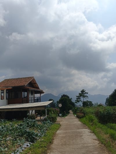 A concrete path stretches into the distance, flanked by lush green vegetation and a cultivated field on the left. A two-story building with a terracotta-tiled roof stands to the left of the path, featuring a wooden porch and a covered outdoor area. Mountains are visible in the background, shrouded in a soft haze. The sky is filled with heavy, overcast clouds, suggesting an impending storm.