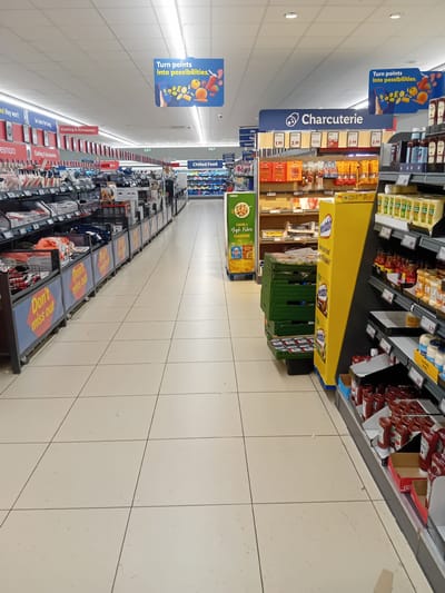 This is a full-shot, eye-level, interior photograph of a supermarket aisle, likely in Inverness, United Kingdom. The aisle is wide and well-lit with artificial ceiling lights. The floor is tiled in a light beige color, with visible tile lines. On the left side of the aisle, shelves are stocked with various non-food items, including clothing and accessories, as indicated by signage. Some of these items are displayed in bins with the repeated text "Don't miss out." Further down the aisle, more shelves are visible, with products that appear to be packaged food items.
On the right side of the aisle, there is a display of charcuterie products, indicated by a large blue sign with the word "Charcuterie" and an illustration of a pig. Below this sign are shelves stocked with packaged meats and sausages, along with price tags. Further down the aisle on the right, a prominent yellow display stand for "Weetabix" cereal is positioned. Adjacent to the Weetabix display are shelves stocked with bottles of ketchup and other condiments, also with visible price tags. In the background, the aisle continues, with more shelving and products, including what appears to be a chilled food section. Several promotional signs are hanging from the ceiling, including one with the text "Turn points into possibilities." There are no people visible in the image. The overall impression is one of a well-stocked and organized retail environment.
