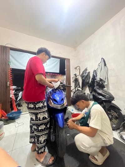 Two individuals are working on motorcycles inside a garage. The individual on the left, wearing a red t-shirt and patterned pants, is standing and appears to be cleaning the front of a blue motorcycle. The individual on the right, wearing a white t-shirt and light-colored pants, is crouched down and appears to be working on the front wheel of the blue motorcycle. A black motorcycle is parked behind the blue one. Various motorcycle parts are visible on the wall and floor. The scene is dimly lit, with fluorescent lights on the ceiling. The location is identified as a garage in Ciakar, Indonesia.
