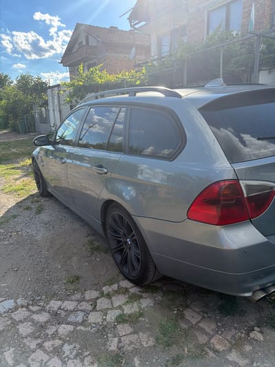 A silver BMW station wagon is parked on a dirt and cobblestone path next to a building with a red-tiled roof. The car is viewed from the rear passenger side, with its tail light and rear wheel clearly visible. The vehicle appears to be in good condition. Behind the car, a brick building with several windows is visible, partially covered by green foliage. The sky above is a vibrant blue with scattered white clouds, indicating it is daytime. The scene suggests a residential or rural setting. There are no people visible in the image.
