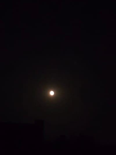 A full moon is visible in a dark night sky over Kostelec na Hané, Czechia. The moon is the brightest object in the image, appearing as a glowing orb against the black backdrop. Below the moon, the silhouette of a building or structure is barely discernible, adding a subtle hint of the urban environment. The overall impression is one of a serene and quiet night.