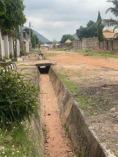 The image depicts a street scene in Kporva, Nigeria. A concrete drainage ditch runs along the side of a dirt road, which appears to be the main thoroughfare. The ditch is partially covered by a concrete slab in the foreground. Lush green vegetation grows along the edges of the ditch and on the roadside. In the background, there are residential buildings with corrugated iron roofs and walls made of concrete blocks and fencing. Power lines are visible overhead, supported by utility poles. A hilly, tree-covered landscape forms the backdrop under an overcast sky. Two individuals are walking in the distance on the dirt road, and a few trees line the street. The overall impression is that of a moderately developed urban or semi-urban area.