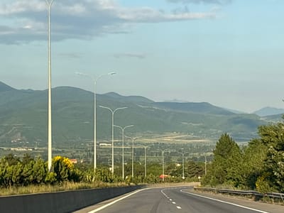 The image depicts a highway curving to the right, flanked by a concrete barrier and a guardrail. A series of tall, modern streetlights line the left side of the road, with some in the foreground and others receding into the distance. Beyond the streetlights, a verdant landscape unfolds, featuring rolling green hills and distant mountains under a hazy sky with scattered clouds. In the middle ground, there is a glimpse of a village or town with buildings nestled amongst trees, and a patch of bright yellow flowering bushes. The scene appears to be captured during daylight, likely late afternoon or early evening, given the soft lighting. The location is identified as the city of Urbnisi, Georgia.