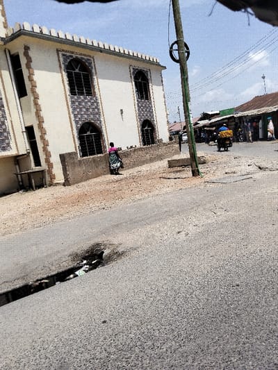 This image depicts a street scene in Mkomani ward, Kenya, under clear daytime conditions.
In the foreground, a paved road surface shows wear and contains a significant linear defect or pothole, approximately 0.5 meters in length, filled with visible debris including light-colored plastic material.
To the left, a multi-story building with a light-colored exterior features several arched windows fitted with dark grilles, framed by decorative, textured brickwork. The building's roofline is adorned with a crenellated parapet. A low concrete wall delineates the building's property from an unpaved, dusty shoulder running parallel to the road. A person, dressed in a pink top and a green and black patterned lower garment, walks away from the camera along this dirt shoulder.
In the mid-ground, slightly right of center, a tall wooden utility pole stands upright, supporting multiple overhead electrical wires and featuring a dark circular object affixed near its top.
Further in the background, to the right, a row of single-story structures with corrugated metal roofs lines what appears to be another street or alley, indicative of a commercial area. A person on a motorcycle, wearing a blue shirt, is visible on this background street, transporting a large yellow bundle on the vehicle's rear. Other indistinct figures are present in the background. The sky above is light blue with sparse white clouds.
