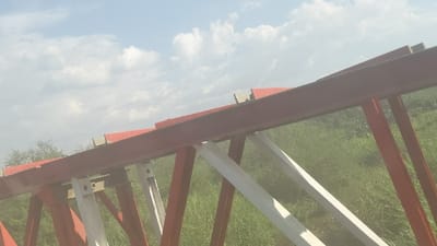 An angled view showcases a section of a bridge structure against a backdrop of sky and foliage. The bridge features a prominent red horizontal beam, supported by alternating red and white diagonal supports. The sky overhead is light blue with scattered white clouds. Lush green grass and vegetation fill the lower portion of the frame, softening the architectural lines.