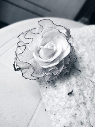The image displays a monochrome, close-up view of a decorative artificial flower, identified as a white rose bud. The central rose bud appears to be crafted from a smooth, matte material. It is surrounded by ruffled, translucent fabric petals, likely organza or similar material, which feature a dark, glitter-lined trim along their scalloped edges. A dark-colored clip, possibly a hair clip or brooch pin, is partially visible on the left side, attached to the base of the flower.

The artificial flower rests on a surface composed of two distinct materials. The right and upper portions of the surface are covered by a light-colored, textured, fibrous material, resembling a blanket, towel, or carpet, which exhibits scattered dark flecks or debris. To the left and underneath the clip, a smoother, lighter-colored surface is visible, possibly a tiled or laminated surface, showing a faint horizontal line. No people, specific actions, or interactions are discernible in the scene. The image is located in Harasta, Syria.